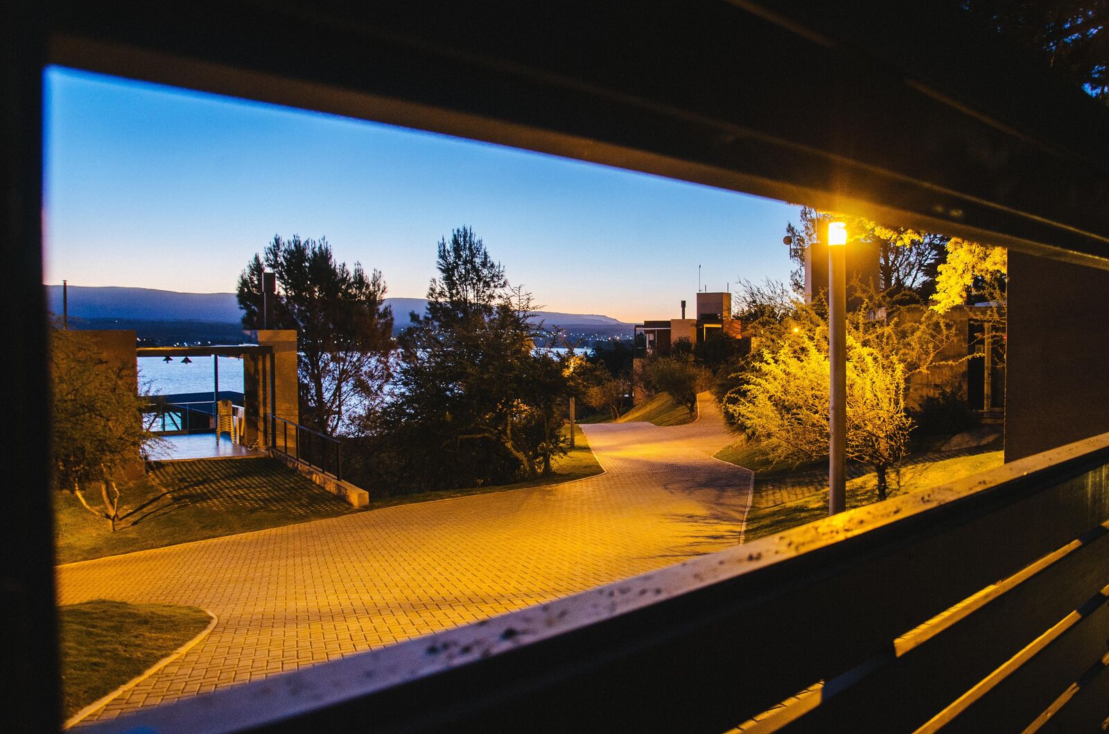 Vista nocturna del camino de acceso iluminado, con el Lago Los Molinos y las luces del pueblo al fondo
