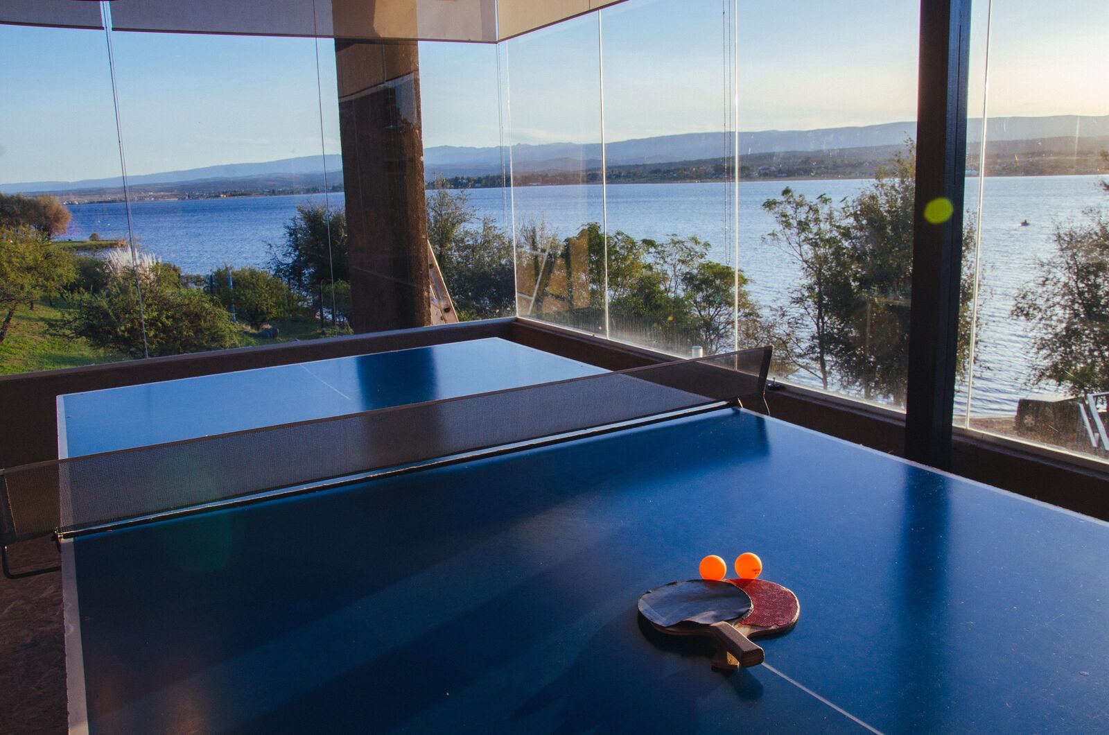 Mesa de ping pong con paletas y pelotas, con el Lago Los Molinos de fondo a través del ventanal